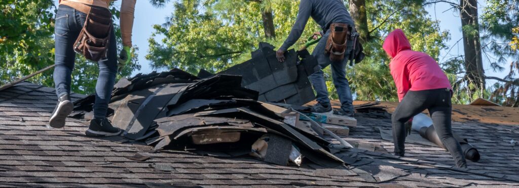 A team of workers removes old shingles from a roof, preparing for replacement amid lush greenery, demonstrating a roof repair and maintenance process.