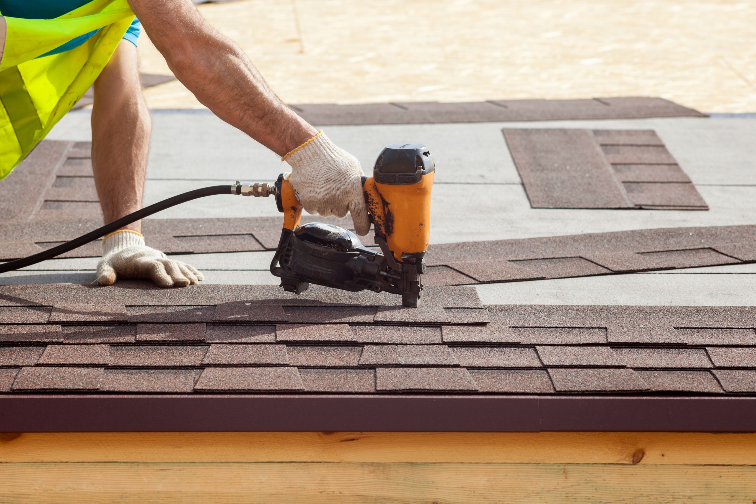 A roofing contractor in a safety vest uses a nail gun to install asphalt shingles on a roof.