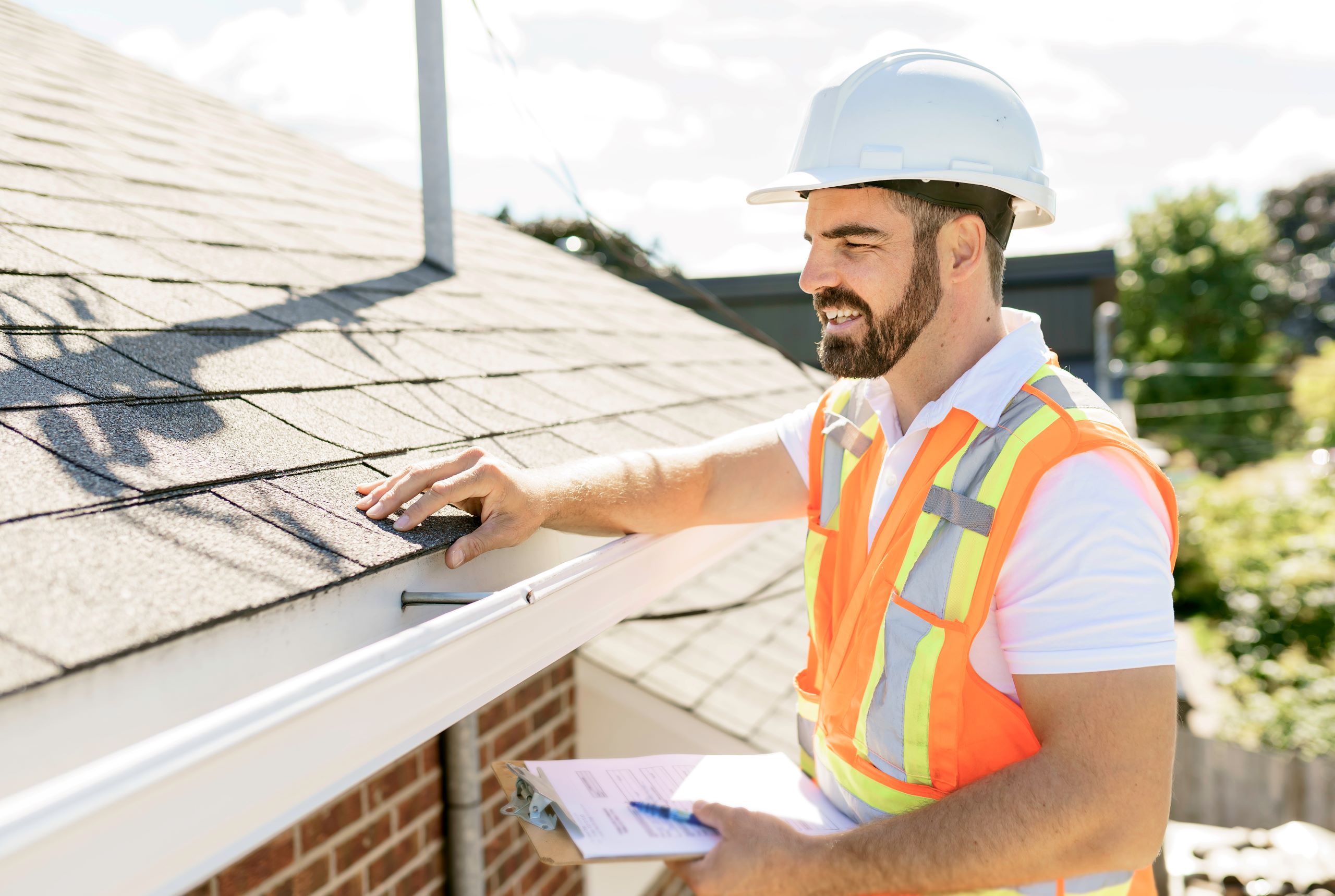 A roofing contractor wearing a safety vest and hard hat inspecting shingles on a residential roof, holding a clipboard for notes.