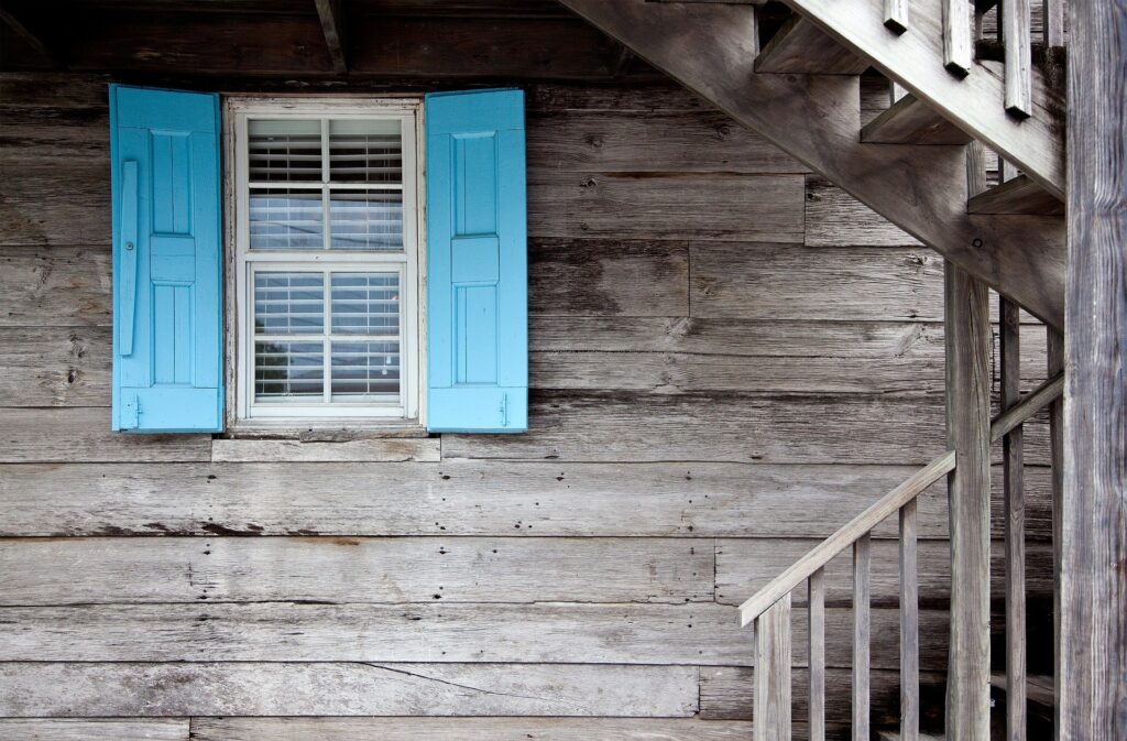 A rustic wooden exterior with weathered wood siding and a single window framed by bright blue shutters, next to a wooden staircase.